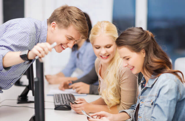 Zwei lächelnde Studentinnen mit Smartphones sitzen in einem Computerraum, ein Student beugt sich über den Bildschirm zu ihnen